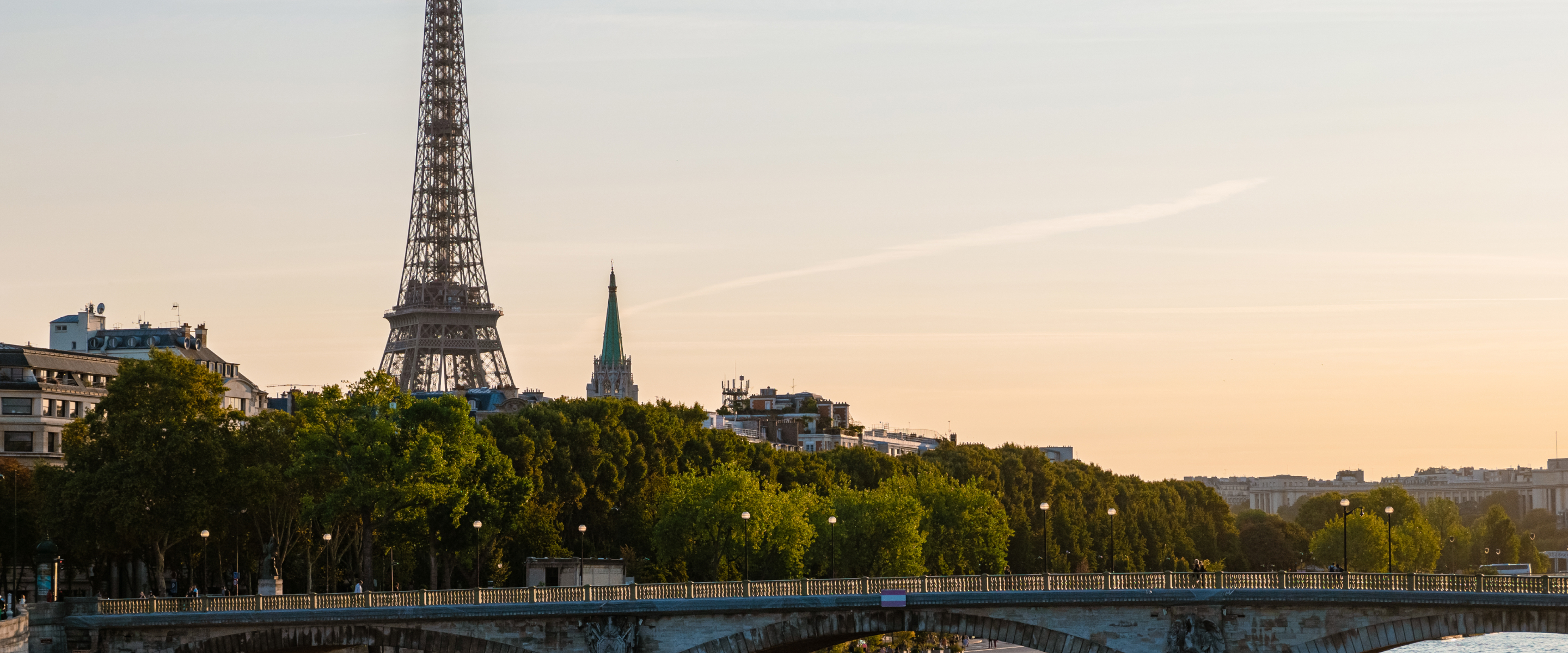 Eiffel Tower rising above the Paris skyline, with a tree-lined bridge over the Seine.