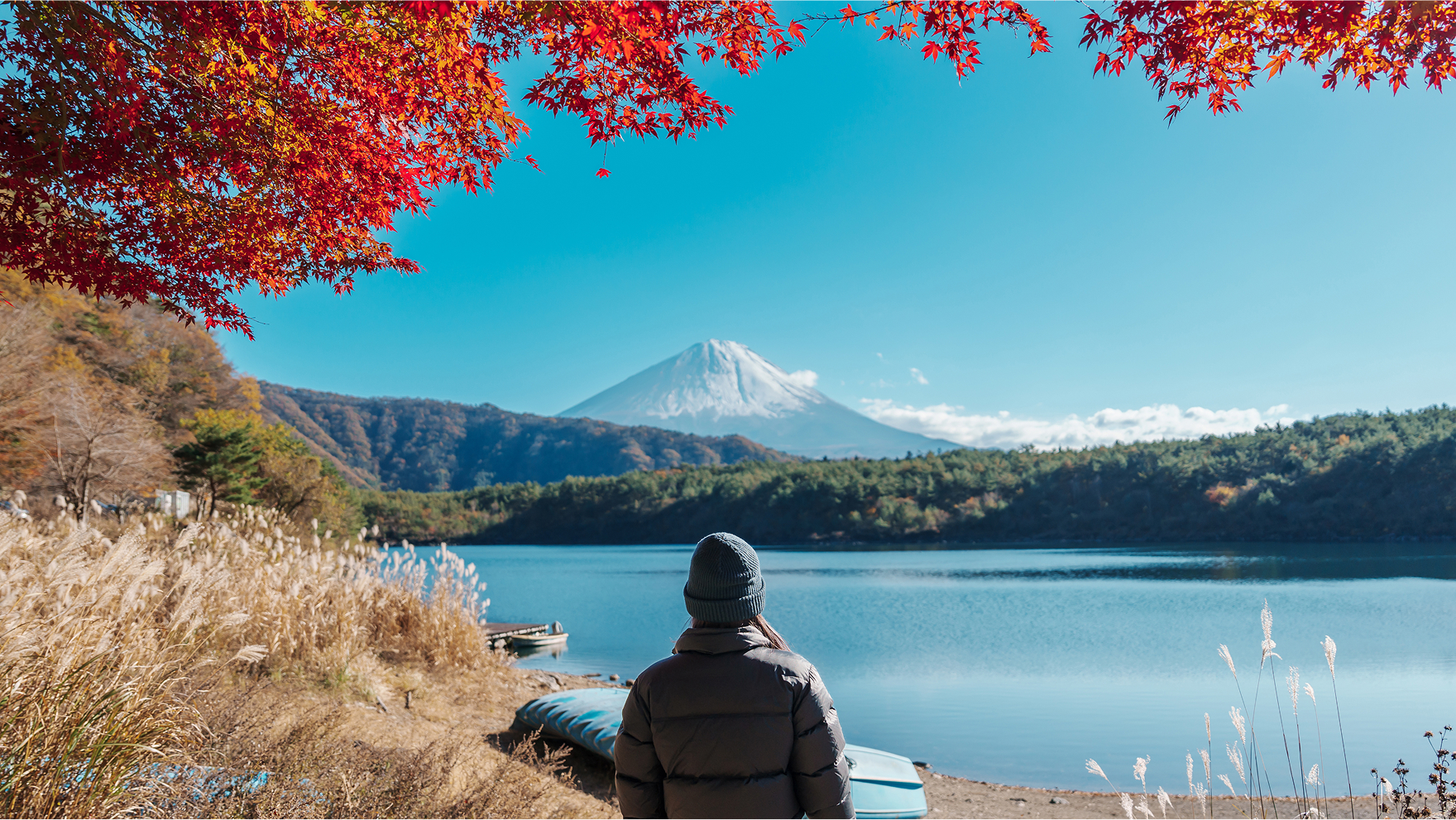 Person in a winter jacket looking at Mount Fuji across a tranquil lake framed by autumn foliage