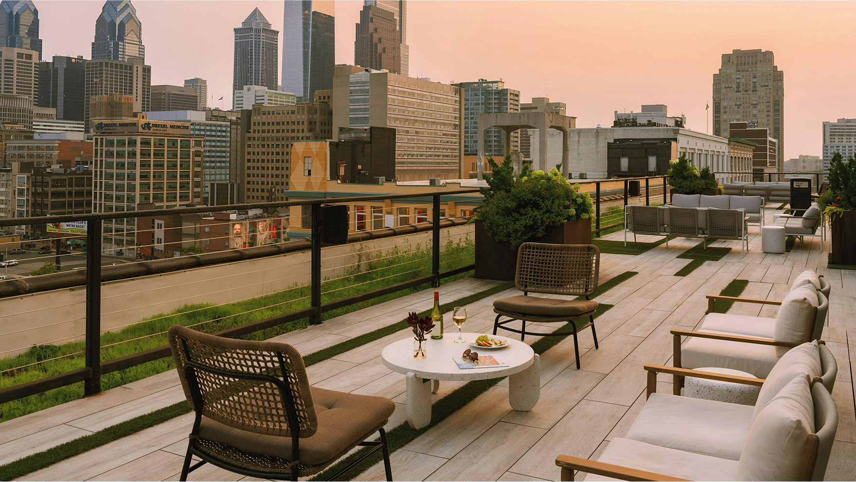  Stylish rooftop lounge area overlooking a city skyline at sunset, featuring modern seating and a table with wine and snacks.