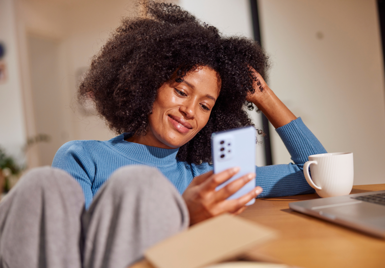 Smiling woman sitting on a couch looking at her phone with a coffee mug and laptop nearby