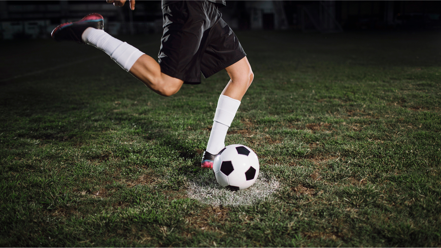 A soccer player in black shorts swinging their leg to kick a soccer ball under stadium lights on a grassy field