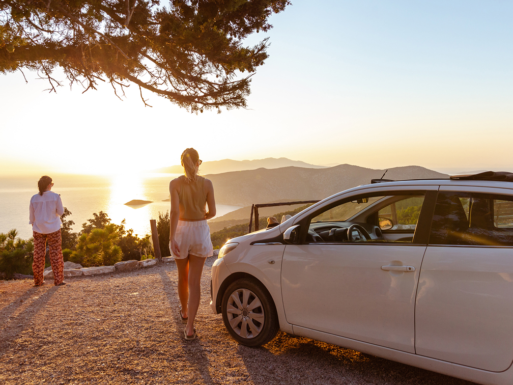 Two people standing next to a white car admiring a scenic sunset view over the ocean from a hilltop overlook