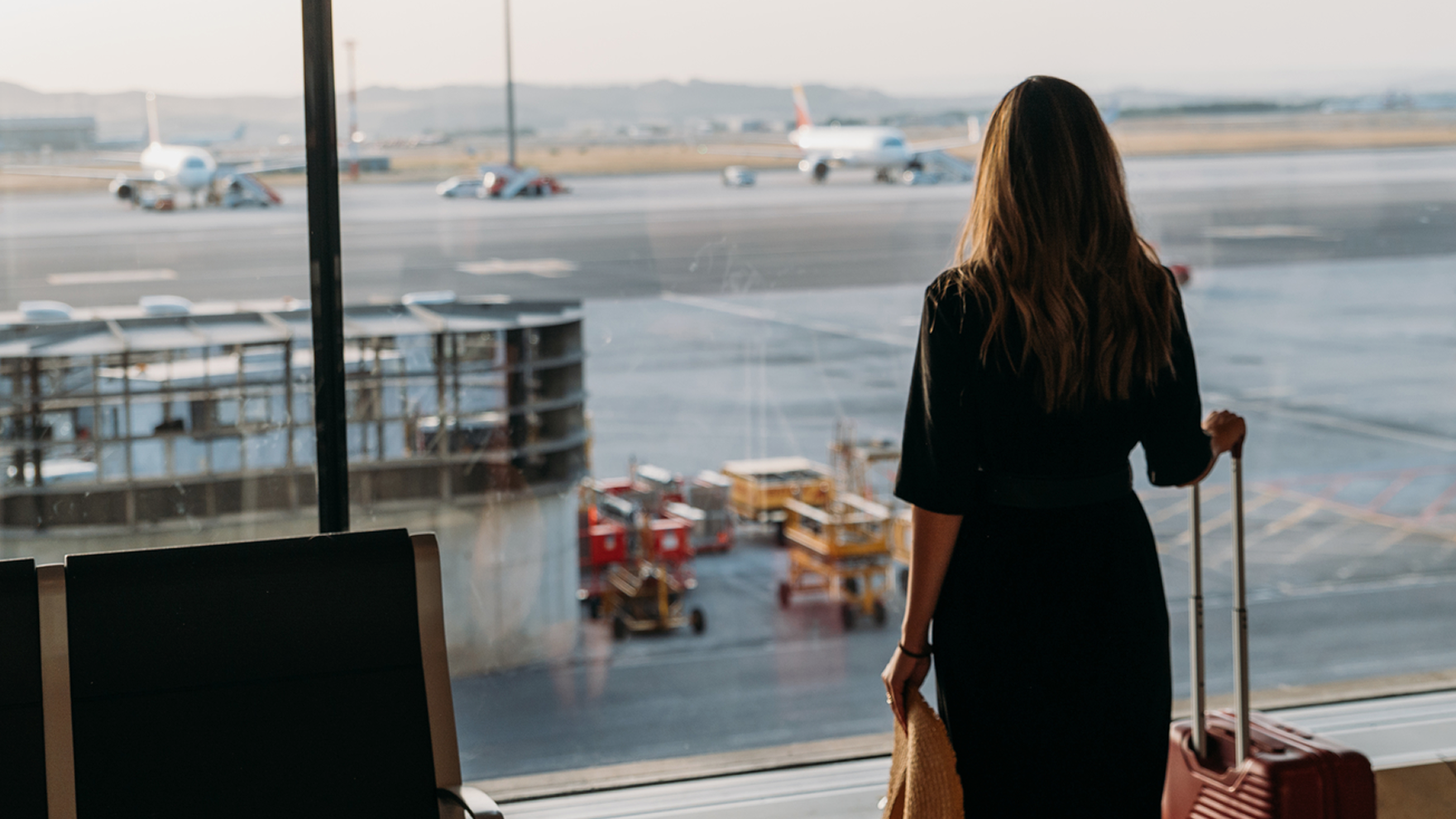 Person standing by an airport window with a rolling suitcase, watching planes on the runway.