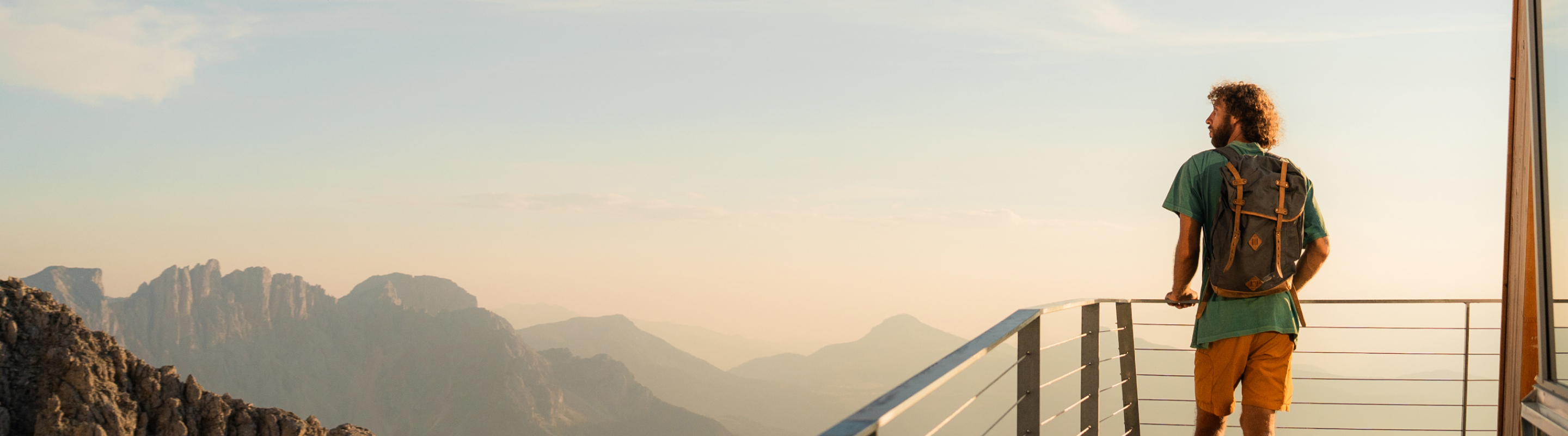Person standing on a viewing platform above a forested valley at sunset, looking toward distant mountains under a colorful sky.