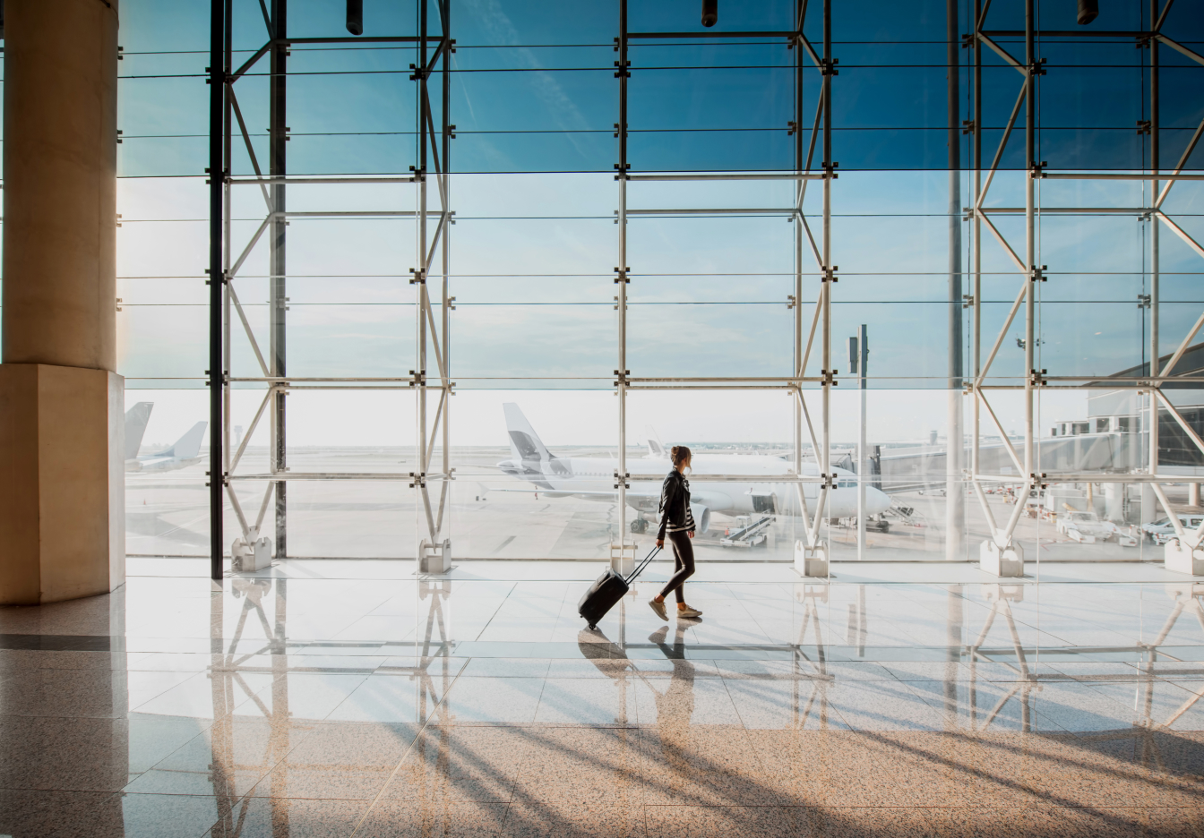 Traveler walking through a bright airport terminal with large windows and a rolling suitcase.