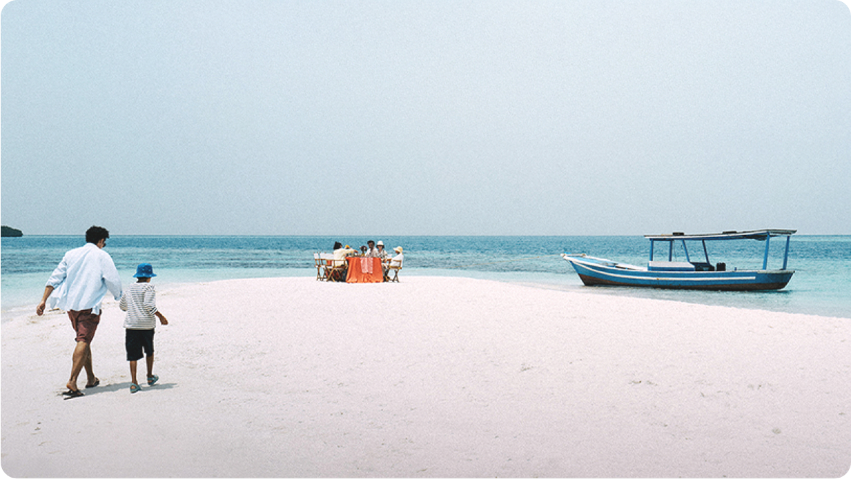 Two people walking on a white sand beach towards a group eating at a dining table set up on the sand, with an island in the distance and a small boat nearby.