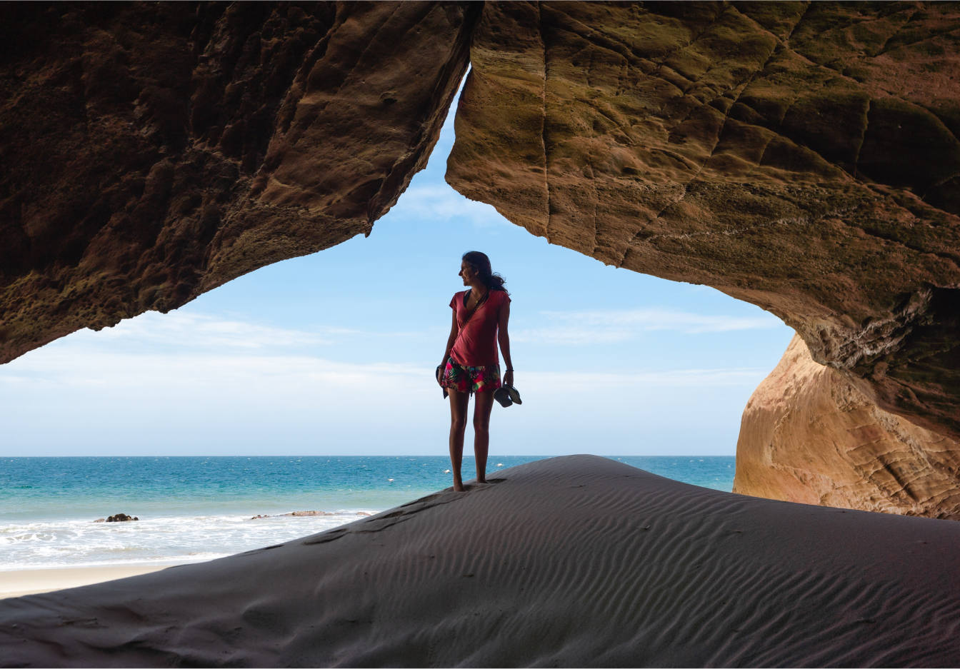 Woman standing silhouetted at the entrance of a sandy beach cave, holding sandals, looking out toward the ocean under a blue sky