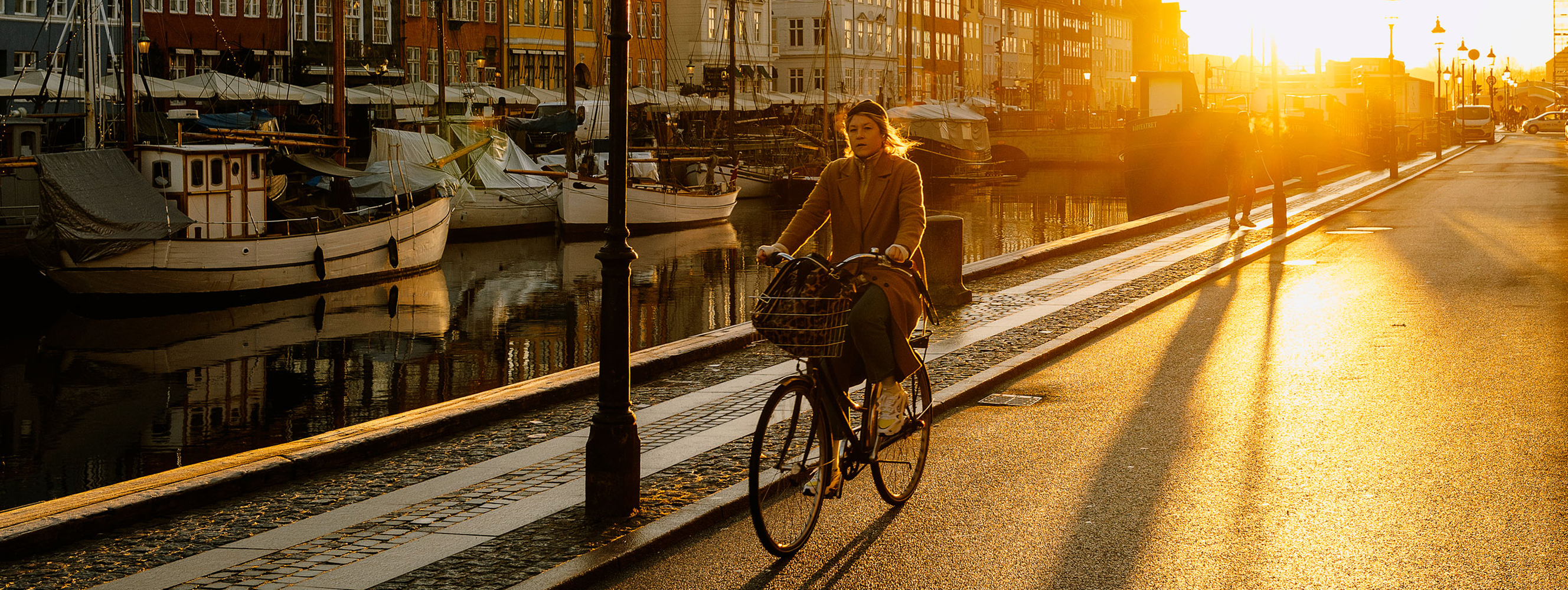 A woman rides a bicycle along a canal in Copenhagen, with boats docked on the water.