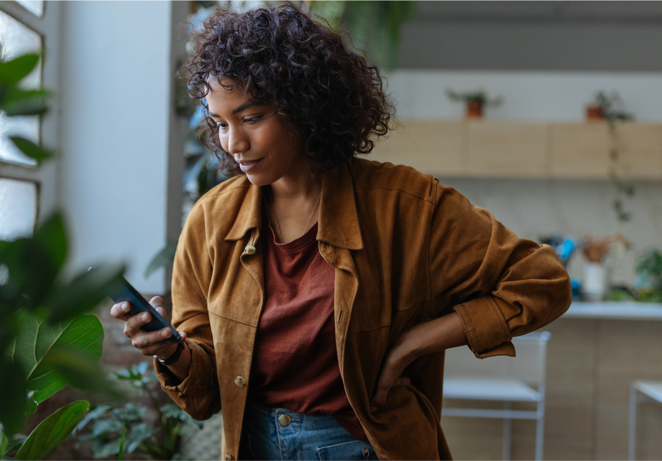 Woman with curly hair in a brown jacket looking at her phone near a bright window with plants