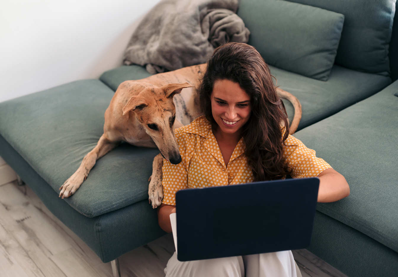Woman on blue sofa with her dog beside her, smiling while using a laptop