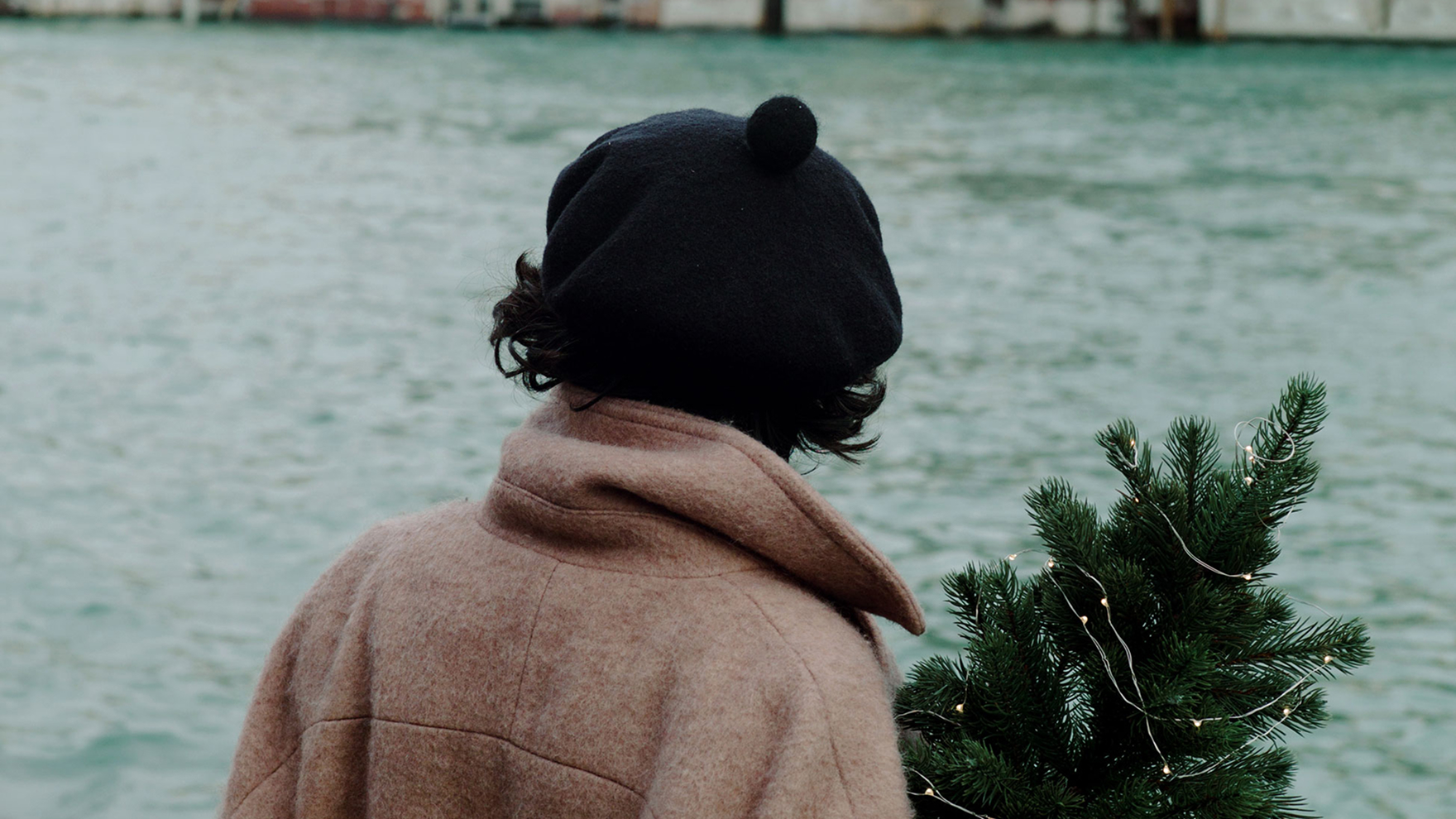 Woman wearing a black beret and tan coat holding a small Christmas tree, viewed from behind at a waterfront.