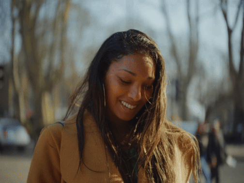 A woman in a bright yellow jacket stands on a city sidewalk, smiling and scrolling on her smartphone. Behind her, pedestrians walk past and cars drive by, both appearing blurred in the background to emphasize her focus and expression.