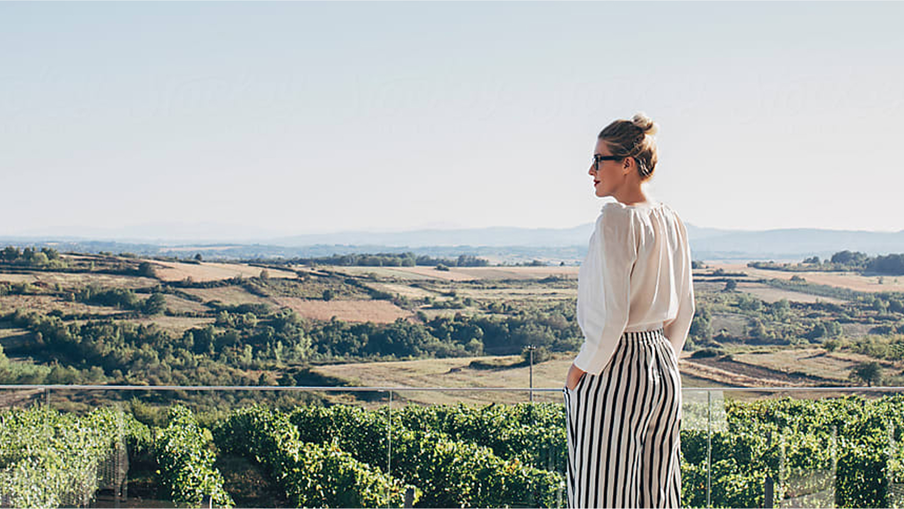 Woman standing on a terrace gazing out over sunlit vineyards and rolling hills under a clear sky.