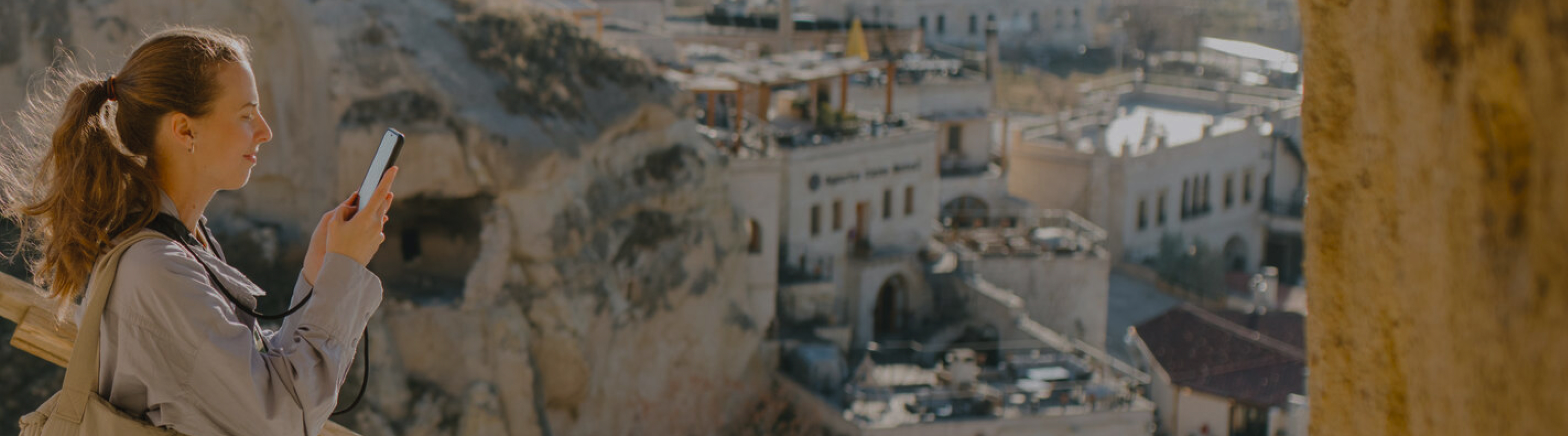 Woman standing outdoors and holding up a smartphone to take a photo, with a rocky cliff face in the foreground and sunlit buildings in the background. The image captures a travel moment in side profile, with warm light, a softly focused cityscape, and the subject’s attention directed toward photographing the view.