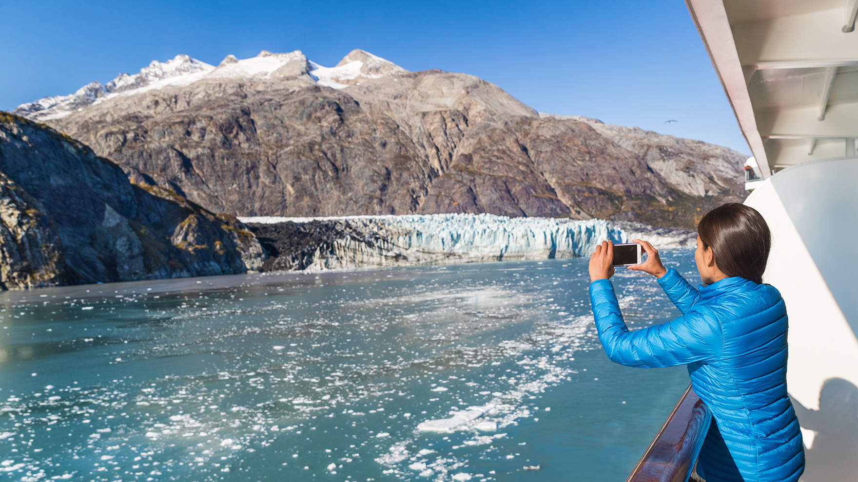 Person in a bright blue puffer jacket standing on the deck of a boat, taking a photo of a glacier and snow-capped mountains across icy blue water under a clear sky.