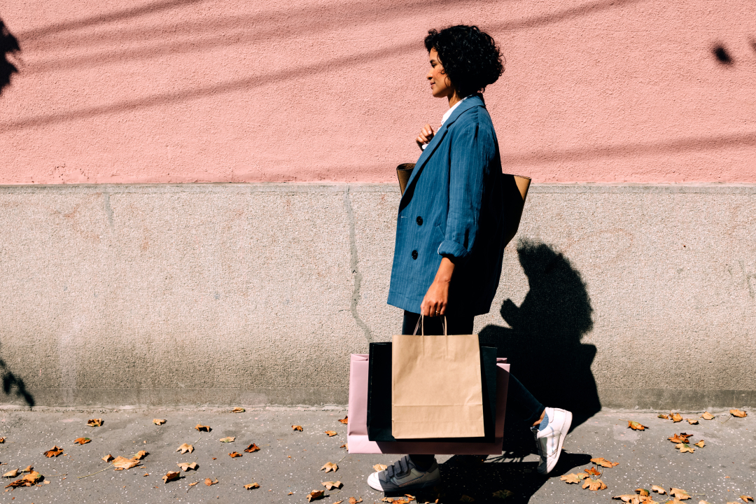 Person walking along a city sidewalk carrying multiple shopping bags against a pink wall.