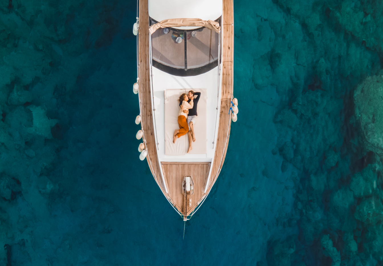 Couple relaxing together on the deck of a yacht, lying side by side and looking up, surrounded by clear blue ocean water.