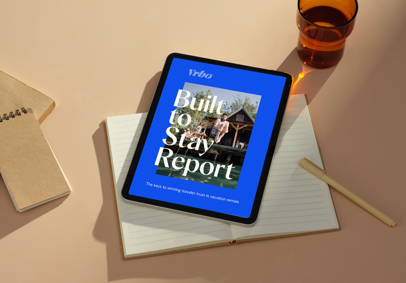 A tablet displaying the Vrbo Built to Stay Report cover rests on top of an open lined notebook, with a pen and glass placed on a softly lit desk around it.
